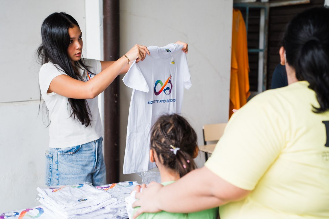 Athalia Suarez holds up a white Kidfinity and Beyond shirt at a distribution table.