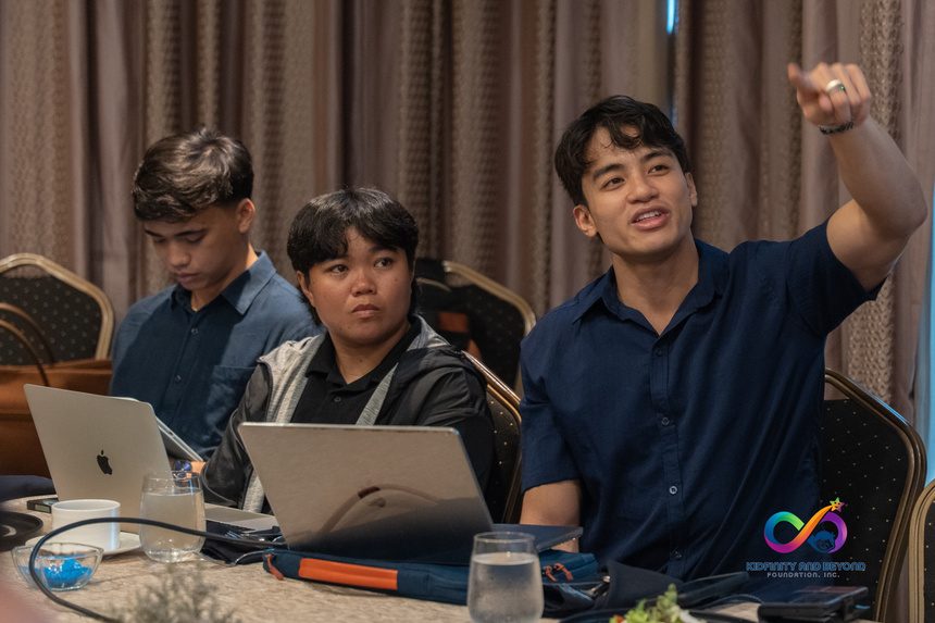 Three men at a table with laptops, one pointing at a presentation.